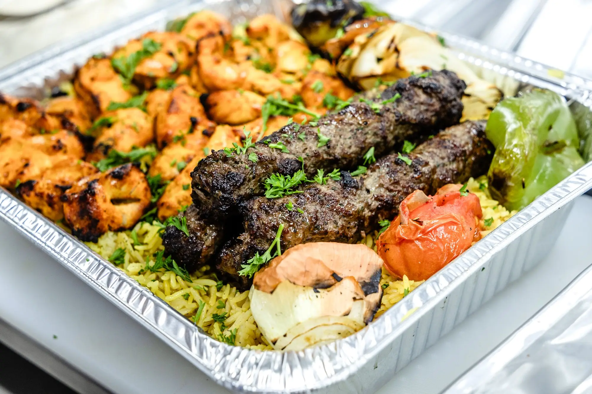 Beef kofta and chicken tawook over yellow rice with grilled tomato and onion, in a catering tray.
